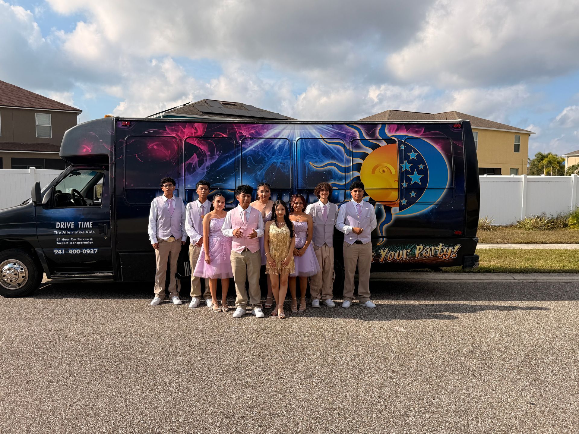 Prom group of teens in pastel formalwear posing in front of a colorful sun-and-moon party bus parked on a suburban street under a cloudy sky