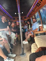 Group of friends relaxing inside a wood-paneled party bus with a center dance pole, blue LED accent lighting, colorful window murals and casual summer outfits.