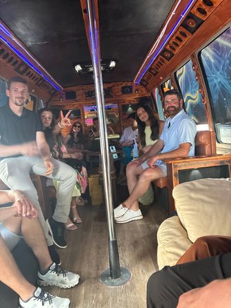Group of friends relaxing inside a wood-paneled party bus with a center dance pole, blue LED accent lighting, colorful window murals and casual summer outfits.