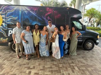 Group of friends in summer dresses and shirts holding drinks, smiling and posing in front of a colorful party shuttle van with a cosmic sun mural and palm trees at a tropical resort entrance