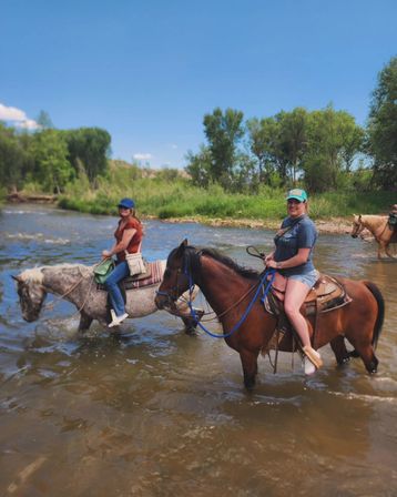 Two riders on horseback crossing a shallow river on a sunny summer day, scenic grassy riverbank and trees in the background — outdoor horseback riding and river crossing