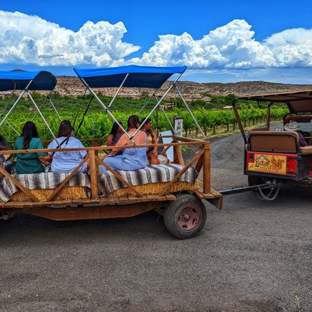 Passengers riding a rustic hay-wagon with blue canopies and striped blankets, towed by an open jeep past green grapevines and rocky mesas under a bright blue sky with dramatic white clouds.
