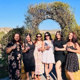 Six friends in summer dresses holding wine glasses under a leafy arch at a sunny outdoor vineyard-like setting, celebrating a bachelorette with rolling hills in the background.
