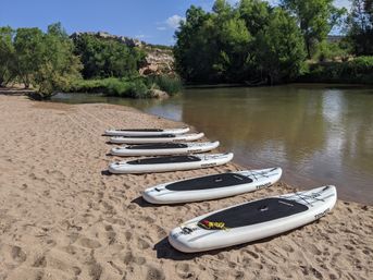 Row of stand-up paddleboards lined up on a sandy riverbank beside a calm, tree-lined river with rocky cliffs and a sunny blue sky.