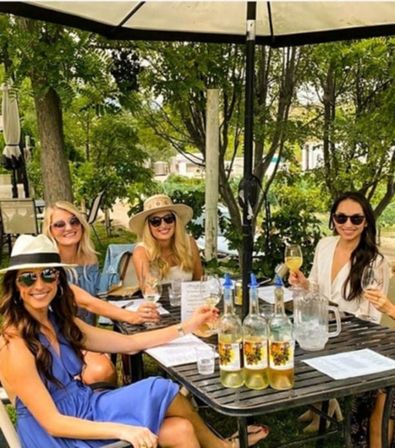 Four friends in summer dresses and sunglasses sipping white wine at a shaded outdoor patio table with bottles, menus and leafy trees in the background.