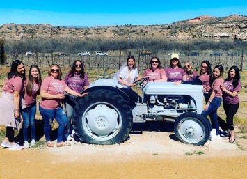 Group of women in matching mauve shirts posing on and around a vintage gray tractor at a sunny vineyard with dry, hilly landscape in the background.