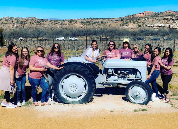 Group of women in matching mauve shirts posing on and around a vintage gray tractor at a sunny vineyard with dry, hilly landscape in the background.