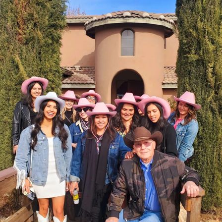 Smiling group in pink cowboy hats and denim with a man in a brown cowboy hat posing on the porch of a Mediterranean-style stucco villa with clay tile roof and tall cypress trees