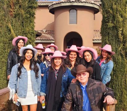 Smiling group in pink cowboy hats and denim with a man in a brown cowboy hat posing on the porch of a Mediterranean-style stucco villa with clay tile roof and tall cypress trees