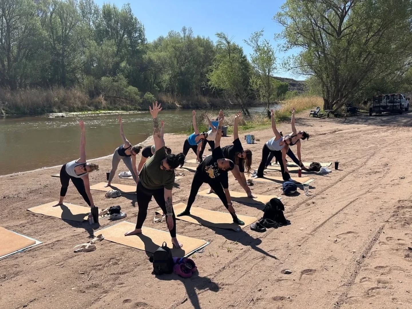Group practicing outdoor yoga on mats along a sandy riverbank, participants stretching in triangle pose under leafy trees on a sunny day