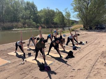 Group practicing outdoor yoga on mats along a sandy riverbank, participants stretching in triangle pose under leafy trees on a sunny day