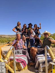 Smiling group of friends on a sunny vineyard hayride, seated on blanket-covered hay bales, holding wine glasses with rolling vineyard hills and a clear blue sky in the background.