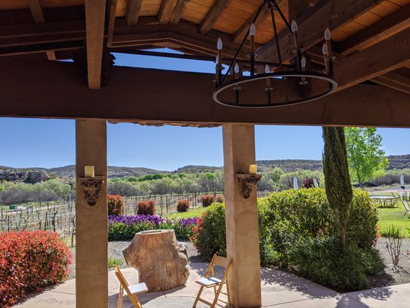 Cozy sunlit rustic patio with wooden beams and chandelier overlooking a vineyard and rolling hills, featuring a tree-stump table, two folding chairs, colorful spring flowers and trimmed bushes.