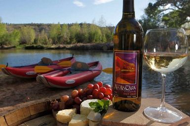 Sunlit riverside picnic with a bottle of white wine and filled glass, cheese and red grapes on a wooden barrel, and red kayaks pulled up on a calm river with a tree-lined shore.