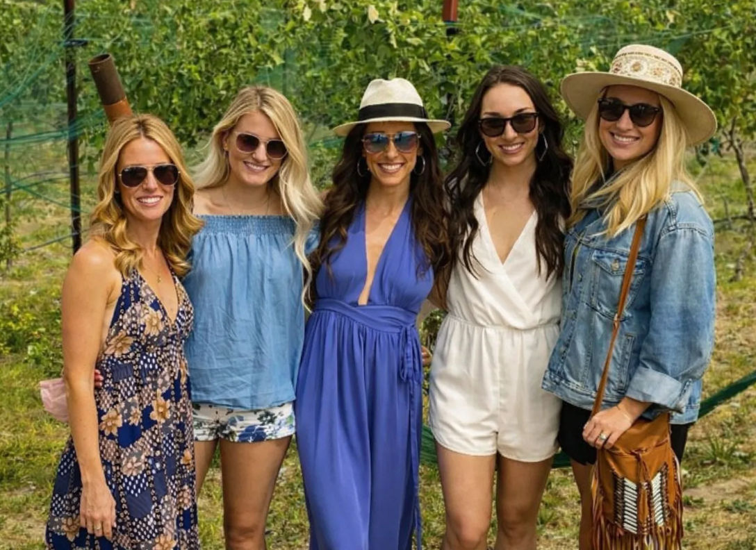 Five women in summer outfits and sunglasses smiling and posing among grapevines at a sunny vineyard — hats, dresses, rompers and a denim jacket for a casual wine-country group photo.