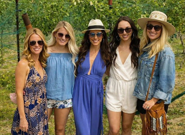 Five women in summer outfits and sunglasses smiling and posing among grapevines at a sunny vineyard — hats, dresses, rompers and a denim jacket for a casual wine-country group photo.