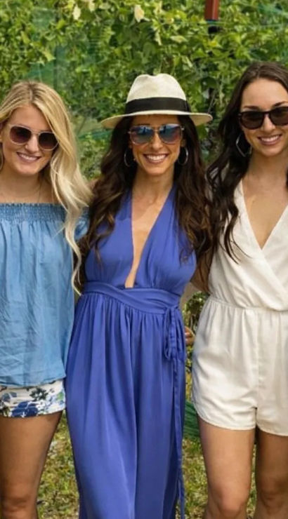 Five women in summer outfits and sunglasses smiling and posing among grapevines at a sunny vineyard — hats, dresses, rompers and a denim jacket for a casual wine-country group photo.