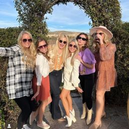 Six women in sunglasses posing under a vine-covered arch in a sunny, hilly outdoor setting, wearing colorful fall outfits and one sipping wine.