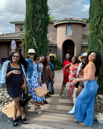 A group of women in colorful summer outfits smiling and posing on a wooden walkway in front of a Mediterranean-style stucco villa with a tiled roof and tall cypress trees.