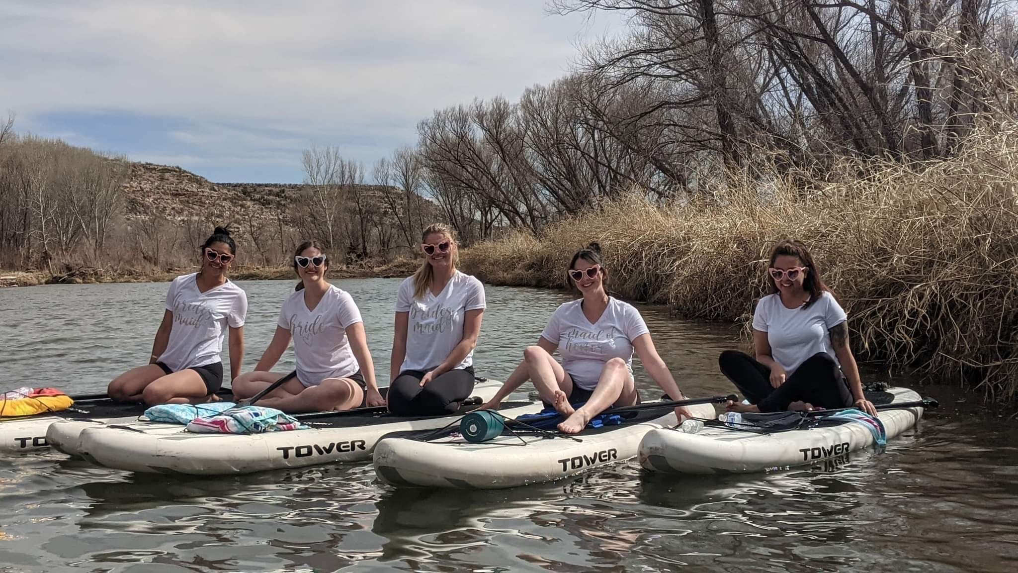 Five friends in matching white shirts and heart-shaped sunglasses seated on paddleboards on a calm river, with bare trees and dry grassy banks under a cloudy spring sky.