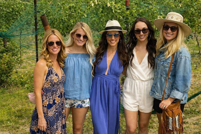 Five women smiling and posing in a sunlit vineyard, wearing summer dresses, rompers, denim jacket, hats and sunglasses for a wine-country outing.