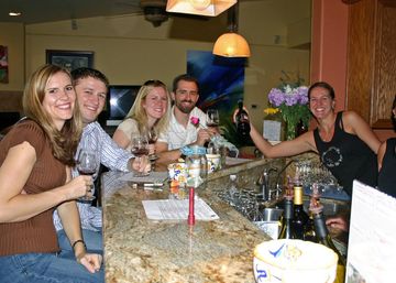 Group of friends enjoying wine tasting at an indoor bar counter, smiling and raising glasses and a bottle over a granite countertop with pendant lights and flowers in the background.