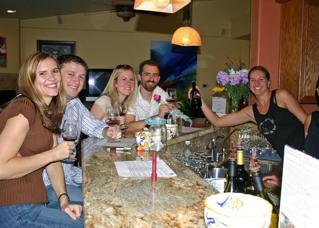 Group of friends enjoying wine tasting at an indoor bar counter, smiling and raising glasses and a bottle over a granite countertop with pendant lights and flowers in the background.