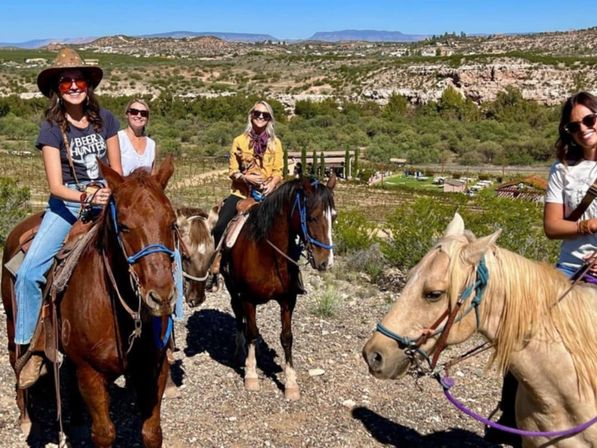 Four riders on a sunny horseback trail ride across arid foothills, mounted on chestnut, bay and palomino horses with canyon mesas and vineyard rows visible below