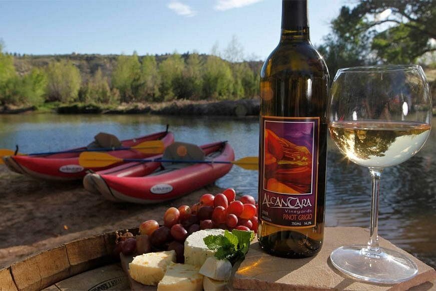 Sunlit riverside picnic with a bottle of white wine and filled glass, cheese and red grapes on a wooden barrel, and red kayaks pulled up on a calm river with a tree-lined shore.