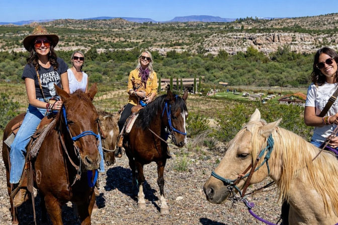 Group of four riders on horseback in a sunny vineyard and arid canyon landscape — chestnut, bay and palomino horses with rolling hills and blue sky in the background.