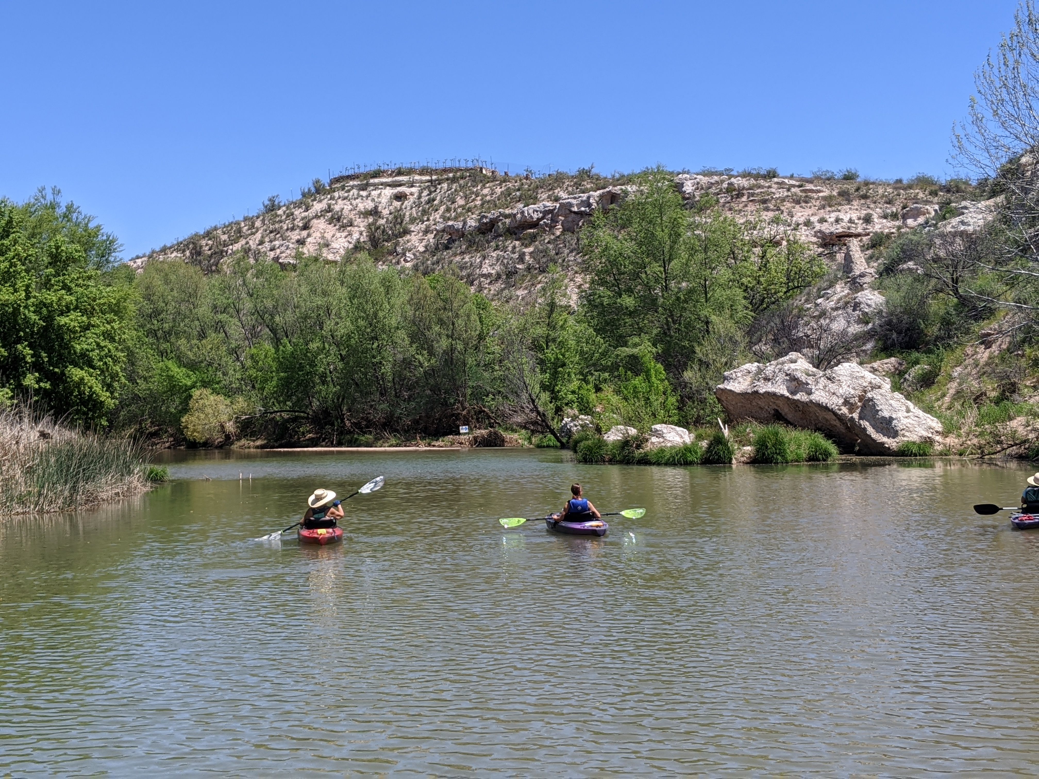Two kayakers gliding across a calm river framed by green trees, reeds and a sunlit rocky hillside under a clear blue sky