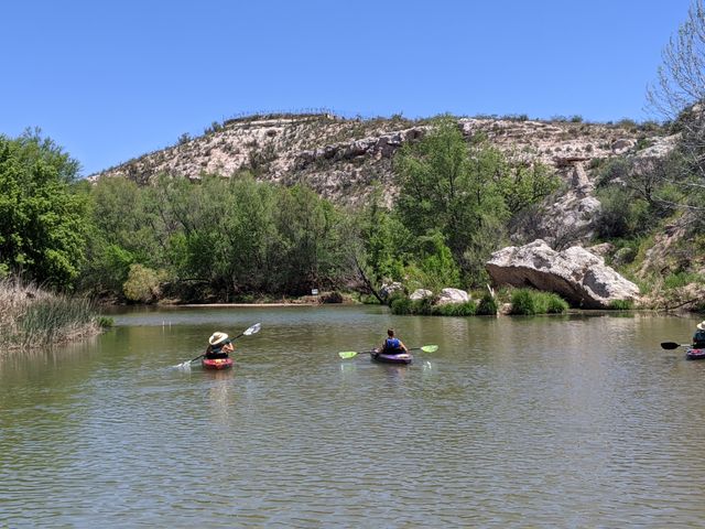Two kayakers gliding across a calm river framed by green trees, reeds and a sunlit rocky hillside under a clear blue sky