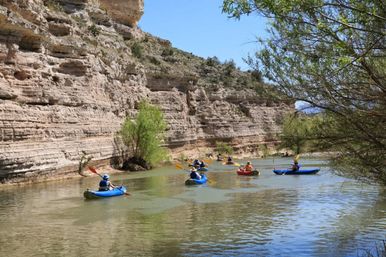 Group of kayakers paddling along a calm river through a sunlit sandstone canyon under a clear blue sky.