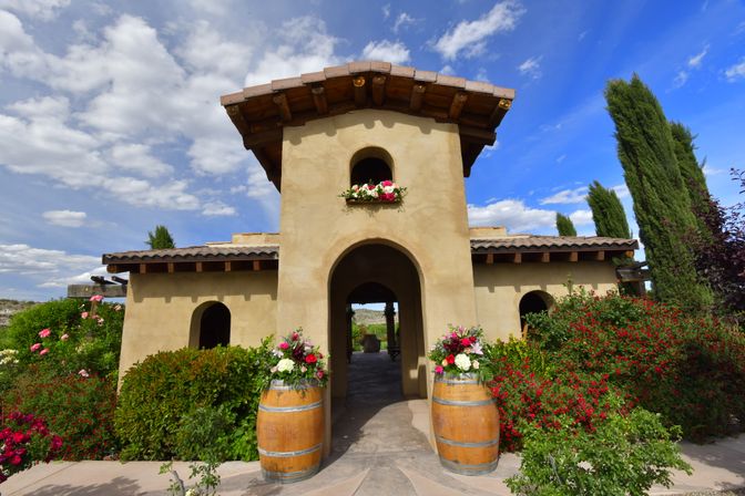 Sunlit Tuscan-style stucco entrance with arched doorway, wine barrels topped with bright floral arrangements, lush rose bushes and tall cypress trees under a blue sky