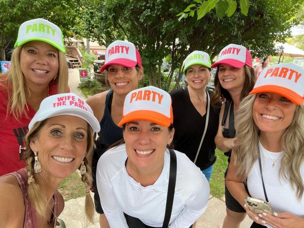 Seven smiling women wearing colorful 'PARTY' trucker hats—one reading 'WIFE OF THE PARTY'—posing for a selfie at a sunny outdoor park/backyard gathering under trees.