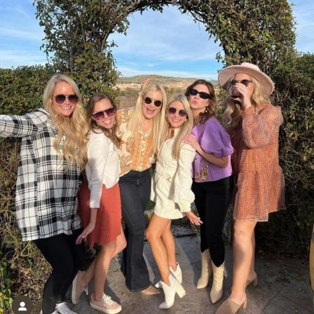 Six women in sunglasses posing under a vine-covered arch in a sunny, hilly outdoor setting, wearing colorful fall outfits and one sipping wine.