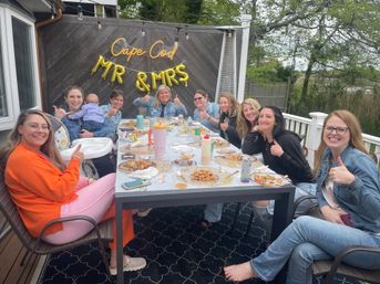 Women gathered on a Cape Cod backyard deck for a bridal shower, giving thumbs up around a long table with plates, a neon "Cape Cod" sign and gold "Mr & Mrs" balloons.