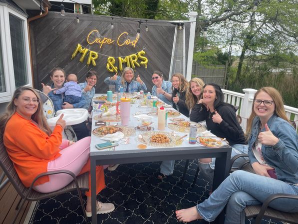 Women gathered on a Cape Cod backyard deck for a bridal shower, giving thumbs up around a long table with plates, a neon "Cape Cod" sign and gold "Mr & Mrs" balloons.