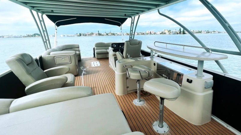 Luxury pontoon boat interior with teak-style deck, plush swivel bar stools, lounge seating and shaded canopy overlooking a calm bay and distant coastal skyline.