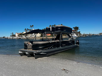 Luxury black pontoon boat beached on a sunny coastal shoreline with palm trees, waterfront homes, docks and a clear blue sky.