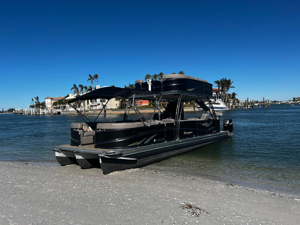Luxury black pontoon boat beached on a sunny coastal shoreline with palm trees, waterfront homes, docks and a clear blue sky.