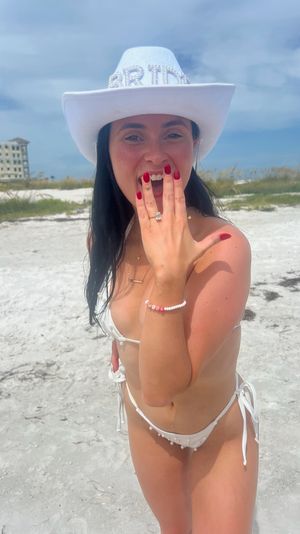 Person in a white bikini and rhinestone "BRIDE" cowboy hat smiling and showing an engagement ring on a sunny white-sand beach with blue sky and beachfront buildings in the background.