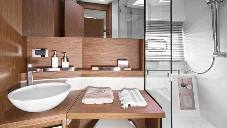 Sleek yacht-style bathroom with wood-accent vanity, white oval vessel sink, folded towels on a wooden shelf, and a glass-enclosed shower with handheld sprayer and porthole-style window.