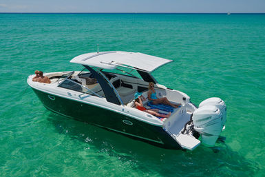 Aerial view of a modern white motorboat with twin outboard engines and a retractable sunshade, passengers in swimsuits lounging on the deck over crystal-clear turquoise tropical water on a sunny day.