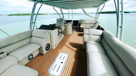 Spacious pontoon boat deck with teak-style flooring and cream leather wraparound seating under a shaded canopy, turquoise coastal water and small islands beyond.