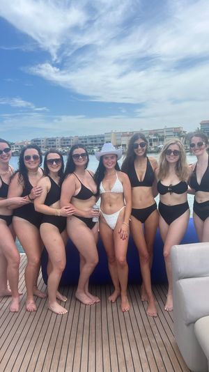 Eight women in swimsuits—seven in black bikinis and one in a white bikini with a white hat—smiling and posing on a yacht deck at a coastal marina with boats and waterfront condos under a bright blue sky.