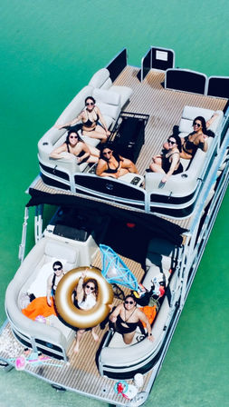 Aerial view of a pontoon boat on turquoise lake water with a group of people in swimsuits lounging on the deck, one holding a gold inflatable ring — summer boating party scene.