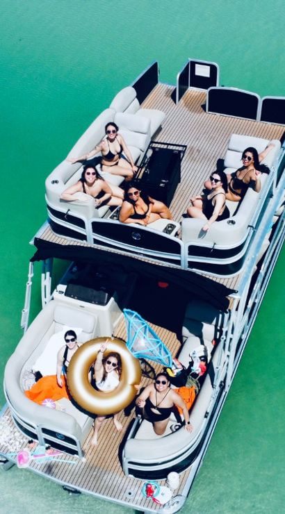 Aerial view of a pontoon boat on turquoise lake water with a group of people in swimsuits lounging on the deck, one holding a gold inflatable ring — summer boating party scene.