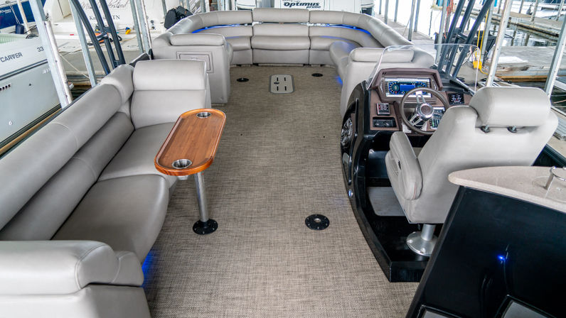Pontoon boat interior docked at a marina with light-gray U-shaped seating, wooden cup-holder table, woven carpet flooring, blue accent lights and a captain’s helm with steering wheel and chair.