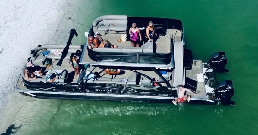 Aerial view of a black double-decker pontoon boat beached on a sandy sandbar in clear emerald-green water with a group of people relaxing and sunbathing.
